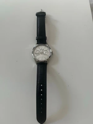 Svart klocka Festina - Stilren klocka från Festina  med svart läderarmband och silverfärgad boett. Urtavlan är vit med silverdetaljer och två mindre urtavlor. Perfekt accessoar för en clean look. Behövs byta batterier