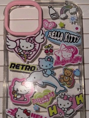 Hello Kitty mobilskal med stickers - Snyggt genomskinligt mobilskal med massor av färgglada Hello Kitty-stickers, bland annat med delfin, nalle, körsbär och retrotext. Skalets kant runt kameran är rosa. Perfekt för dig som gillar kawaii och vill sticka ut med din mobil!