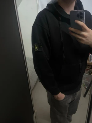 Svart Zip från Stone Island - Säljer en svart hoodie från Stone Island med klassisk logotyp-patch på vänster ärm. Hoodien har huva med dragsko och en stor magficka framtill. Perfekt för dig som gillar streetwear och vill ha en stilren look.