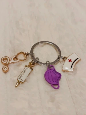 Nyckelring med vårdtema och berlocker - Cool nyckelring med berlocker i form av stetoskop, spruta, munskydd och sjuksköterskehatt. Färgerna är guld, vitt, lila och rött. Perfekt accessoar för dig som gillar vård eller vill ha något unikt på väskan eller nycklarna.