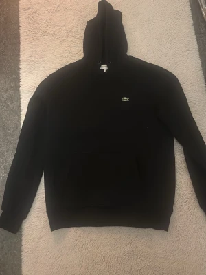Svart hoodie från Lacoste - Säljer en svart hoodie från Lacoste med en skön passform knappt använd säljer pga inte riktigt min stil.