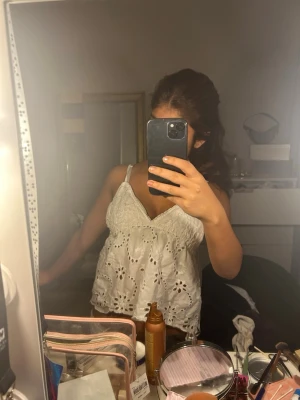 Vit spetsig linnetopp med broderi - Säljer en vit linnetopp med broderade detaljer och spets längs v-ringningen. Toppen är ärmlös med smala axelband och har en luftig, loose passform. Perfekt för sommarens varma dagar och ger en fräsch look.