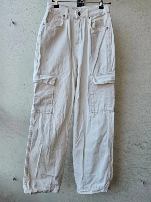 Vita cargobyxor med raka ben - Säljer ett par vita/ljust cremefärgade cargobyxor med hög midja, raka ben och stora fickor på sidorna. Lite stretchigt material. Storlek 34. En liten fläck på framsidan av vänster ben och en liten fläck vid ena bakfickan som man ser närmre på sista bilderna. Byxbenen är slitna längst nere.