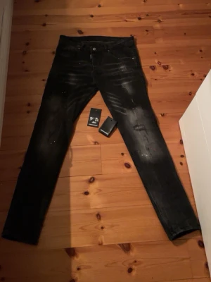 Svarta jeans från Dsquared2 - Säljer ett par svarta jeans från Dsquared2 med slitningar och färgstänk för en edgy look. Jeansen har klassisk femficksmodell, knappgylf och raka ben. Materialet är denim med stretch för extra komfort. Perfekta för dig som gillar streetwear och vill sticka ut.