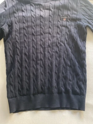 Mörkblå kabelstickad tröja GANT - Kabelstickad tröja i mörkblått från GANT med broderad logga på bröstet. Tröjan är gjord i premium bomull och har ribbade muddar vid ärmslut och nederkant. Klassisk rund halsringning och snygg struktur.