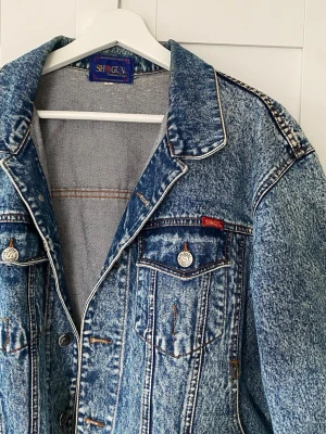 Blå vintage jeansjacka från Shotgun - Säljer en blå vintage jeansjacka från Shotgun med klassisk tvättad look och kontrastsömmar. Jackan har två bröstfickor med lock och silverfärgade knappar med logga. Oversized passform och retrostil, perfekt till våren! Storlek L men jag som är S har haft den som lite oversize. Liten gul rand längs med kragen, men inget man tänker på när man har den på. Se sista bild.