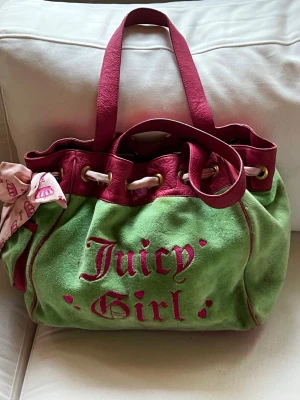 Juicy Couture grön handväska - Grön och rosa handväska från Juicy Couture i mjuk mocka med texten 'Juicy Girl' broderat framtill. Väskan har rosa detaljer, dubbla handtag, snörning upptill och en söt scarf knuten på sidan. Insidan har hjärtformad detalj och tryckt foder.