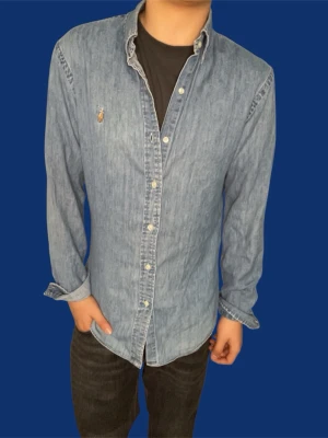 Skjorta från Ralph Lauren - En blå jeansskjorta från Ralph Lauren med buttondown krage och broderad logga på bröstet. Slim fit och knappar framtill och är tillverkad i mjuk bomull. Perfekt för en avslappnad men ändå stilren look ett litet hål vid baksidan