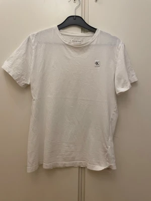 Vit Calvin Klein T-shirt - Vit t-shirt från Calvin Klein med liten CK-logga på bröstet. Klassisk rund halsringning och korta ärmar. Storlek 14år/164cm. Skriv gärna vid minsta fundering!