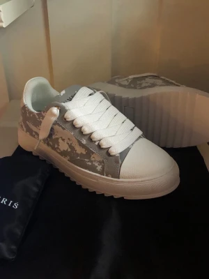 Gråvita sneakers från Numeris - Snygga sneakers från Numeris i grått och vitt med camomönster. Skorna har tjock, räfflad sula och breda vita skosnören. Ovandelen är i textil och syntet, och designen är modern och streetinspirerad. Perfekta för dig som gillar unika detaljer och trendig look.