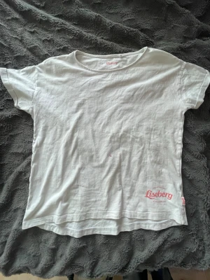 Vit Liseberg t-shirt med tryck - Vit t-shirt från Liseberg med rött tryck på framsidan där det står 'The heart of Liseberg Gothenburg Est. 1923'. Klassisk rund hals och korta ärmar. Perfekt för dig som gillar Göteborgs nöjespark och vill ha en enkel, stilren look.