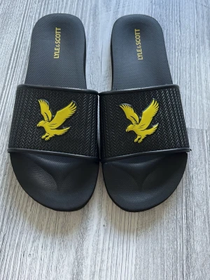 Svarta slides från Lyle & Scott - Säljer ett par svarta slides från Lyle & Scott med gul logga i form av en fågel på ovandelen. Sandalerna har en bred rem med fiskbensmönster och är tillverkade i syntetmaterial. Perfekta för sommaren eller att slappa i hemma.