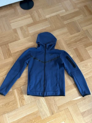 Blå Nike tech hoodie - Snygg blå hoodie från Nike med hel dragkedja framtill och huva. Har en diskret svart Nike-logga på bröstet och en svart detalj över bröstet. Ficka med dragkedja på ena ärmen och två sidofickor. Perfekt för en sportig och avslappnad stil. Har även matchande byxor på min profil:)