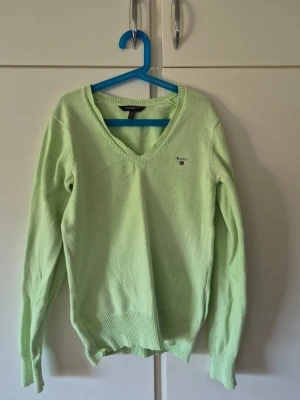Ljusgrön stickad tröja från GANT - Ljusgrön stickad tröja från GANT med V-ringning och diskret logotyp broderad på bröstet. Tröjan har långa ärmar och ribbstickade muddar vid ärmslut och nederkant. Perfekt för dig som gillar klassisk och stilren design. Säljer då den inte används då jag inte är ett fan av färgen längre. Köpt för 1200kr