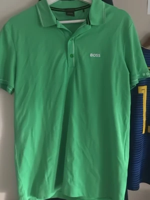 Grön piké från BOSS med vit logga - Snygg grön pikétröja från BOSS med vit logga på bröstet. Slim fit-modell med korta ärmar, krage och knappar framtill. Materialet är troligtvis bomull eller polyester, perfekt för en fräsch och sportig look. Köpt för 799kr