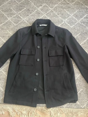 Svart overshirt från Zara - Stilren svart overshirt från Zara med klassisk krage och fyra fickor framtill, två med lock och två snedställda. Jackan har raka ärmar och knäpps med svarta knappar. Perfekt för lager-på-lager och enkel att matcha med olika outfits.