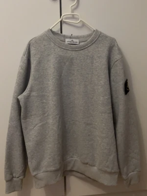 Grå sweatshirt från Stone Island - Snygg grå sweatshirt från Stone Island med klassisk rund hals och ribbade muddar. Ikonisk svart och gul patch på vänster ärm. Perfekt för dig som gillar streetwear och enkel stil. Materialet är mjukt och bekvämt, troligen bomull. Storlek S (170-179) kom jätte gärna med bud 
