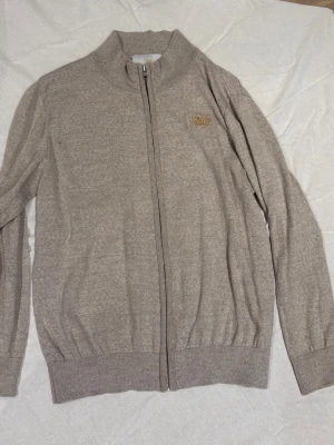 Beige ziptröja med broderad logga - Snygg beige ziptröja från Mellerud med broderad guldfärgad logga på bröstet. Tröjan har hög krage, långa ärmar och ribbade muddar vid ärmslut och nederkant. Perfekt för en stilren och avslappnad look.