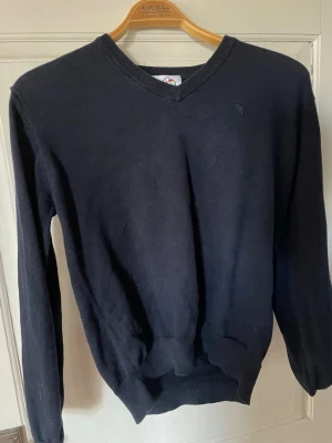 Mörkblå v-ringad tröja - Klassisk mörkblå v-ringad tröja med ribbade muddar vid ärmslut och nederkant. Tröjan har lång ärm och en liten diskret logga på bröstet. Perfekt att matcha med skjorta eller t-shirt under för en clean look.
