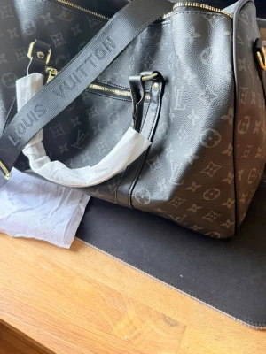 Svart monogramväska - Snygg svart väska  med klassiskt monogrammönster i grått. Väskan har guldfärgade metalldetaljer, dragkedja och både axelrem och handtag. Tillverkad i tåligt lädermaterial och har en rymlig insida med innerfack. Ej äkta 