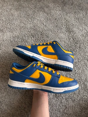 Nike Dunk Low  Ucla blå/gul sneakers - Nike Dunk Low sneakers i blått och gult läder med vit mellansula och blå yttersula. Klassisk låg siluett med perforerad tåbox, gula skosnören och tydlig Swoosh-logga på sidan. Perfekta för dig som vill sticka ut med färg och stil.