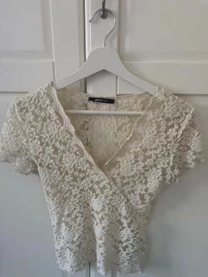 Vit spetsblus från Gina Tricot - Säljer en söt vit spetsblus från Gina Tricot med omlott och v-ringad hals. Blusen har korta ärmar och är helt täckt av blommig spets som ger en romantisk vibe. Perfekt att styla med jeans eller kjol för en trendig look.