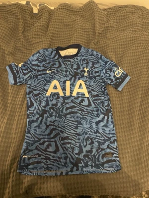 Tottenham bortatröja Nike blå - Säljer en blå Tottenham Hotspur bortatröja från Nike med abstrakt mönster i olika blå nyanser. Tröjan har tryckta loggor, sponsortryck 'AIA' på bröstet och 'KANE 10' på ryggen. Korta ärmar och rund hals. Perfekt för fotbollsfans! Köpte den i Spanien har aldrig använt den 