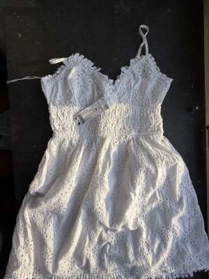 Vit spetsklänning från Gina Tricot - Superfin vit kort klänning från Gina Tricot med broderade spetsdetaljer och smala axelband. Klänningen har v-ringning, smock i midjan och volangkant nertill. Perfekt för sommarens alla tillfällen! Oanvänd!