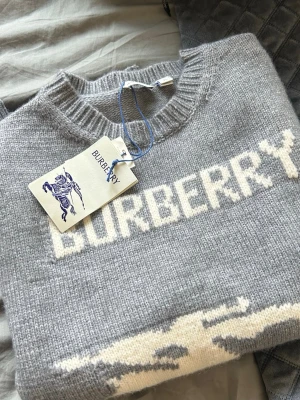 Grå stickad tröja från Burberry - Snygg grå stickad tröja från Burberry med vit logotyp och motiv av riddare framtill. Tröjan har rund halsringning och ribbade muddar. Tillverkad i mjukt material som känns lyxigt och trendigt. Perfekt för dig som vill ha något stilrent med en twist.