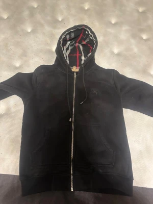 Svart zip-hoodie från Burberry - Snygg svart hoodie från Burberry med dragkedja och klassiskt rutigt foder i huvan. Hoodien har fickor framtill och justerbar dragsko i huvan. Perfekt för dig som gillar exklusiva streetwear-plagg med stilrena detaljer.
