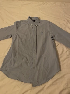 Ljusblå skjorta från Ralph Lauren - Klassisk ljusblå skjorta från Ralph Lauren med button-down krage och broderad logga på bröstet. Skjortan har lång ärm och knäppning framtill. Perfekt för en stilren look och enkel att matcha med jeans eller chinos.