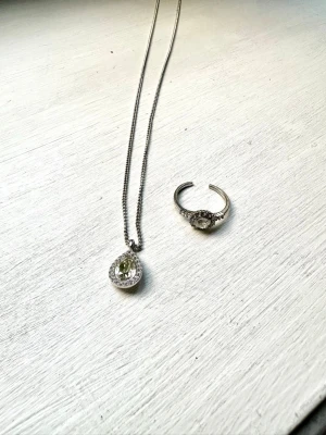 Lily and Rose halsband och ring  - Säljer ett silverfärgat halsband med en droppformad hängande pärla omgiven av små gnistrande stenar. Kedjan är tunn och elegant, perfekt för att ge en stilren touch till din outfit. Märke ej synligt på halsbandet.