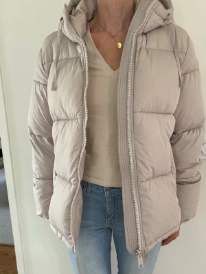 Beige puffer jacka stl XS Nyskick - Beige puffer jacka med huva och dragkedja framtill. Jackan har fickor i sidorna och en dragsko nedtill för justerbar passform. 2 stora fickor på insidan. H&M stl XS, passar en small bra. Använd ett fåtal ggr, är i mkt bra skick, inga defekter. Varm o skön.  Nypris: 499:-