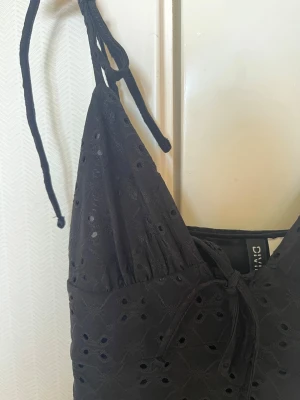 Svart klänning med knytband från Divided - Svart klänning från Divided med knytband på axlarna och broderade hålmönster. Klänningen har en v-ringad front och är ärmlös, perfekt för varma dagar. Materialet är bomull och modellen är en kortklänning med en luftig passform.