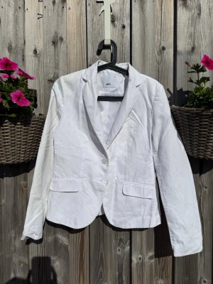Vit kavaj från Gina, storlek 36 - Snygg vit kavaj från Gina i storlek 36. Klassisk modell med två knappar framtill, två fickor med lock och figurnära passform. Perfekt att styla till jeans eller kjol för en clean look. Tillverkad i ett lätt tyg som passar året runt.