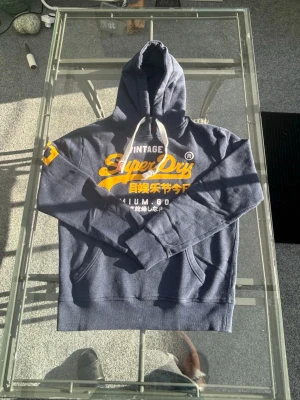 Superdry Hoodie – Navy/Gul – Stl XL - Säljer en klassisk hoodie från Superdry i en snygg marinblå färg med ikonisk gul logotyp och siffran 23 på ärmen. Det här är ett perfekt basplagg för en bekväm streetwear-stil. Tröjan har en skön passform och ett mjukt innerfoder, perfekt att dra på sig till vardags. Kort info:  Märke: Superdry  Modell: Vintage series  Storlek: XL  Färg: Navy (marinblå) med gula detaljer  Skick: Mycket bra skick Skicka ett meddelande om du vill köpa eller har några frågor! 