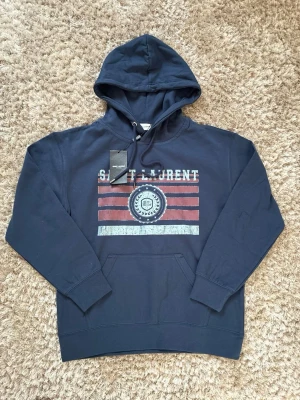 Mörkblå hoodie från Saint Laurent - Snygg mörkblå hoodie från Saint Laurent med tryck i vitt och rött framtill. Klassisk huva med dragsko och stor magficka. Materialet känns mjukt och skönt, perfekt för en avslappnad stil. Märkeslappen sitter kvar.