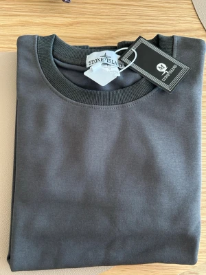 Mörkgrå sweatshirt från Stone Island - Snygg mörkgrå sweatshirt från Stone Island med klassisk rund halsringning och ribbade muddar vid ärmslut och nederkant. Tröjan har en diskret logotyp-patch på ärmen och är tillverkad i mjuk bomull för en skön känsla.