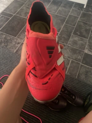 Adidas Predator fotbollsskor röd/vit barn elit - Säljer ett par ikoniska Adidas Predator fotbollsskor i rött och vitt med svarta detaljer. Skorna har klassiska tre ränder på sidan och texten 'PREDATOR' under sulan. Designade för grym kontroll och grepp på planen. Perfekta för dig som vill sticka ut på matchen.