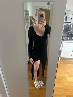 Svart långklänning med slits S-M - Säljer en svart långklänning med lång ärm och djup v-ringning. Klänningen har en hög slits framtill och är figurnära i modellen. Materialet känns stretchigt och mjukt, perfekt för dig som vill ha en stilren och enkel look.