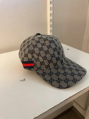 keps med GG-mönster - Säljer en snygg beige keps från Gg med klassiskt blått GG-monogram över hela kepsen. På sidan finns en röd och blå rand som ger extra stilpoäng. Justerbar rem baktill för perfekt passform. Materialet är canvas som känns både lyxigt och tåligt.