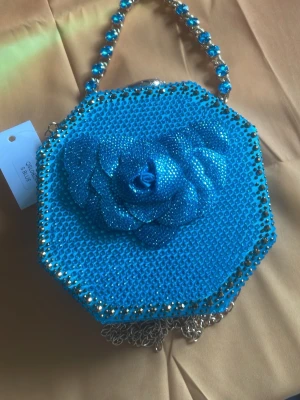 Blå glittrig handväska med blomma - Snygg blå handväska i oktagonform med glittrande stenar och en stor dekorativ blomma på framsidan. Väskan har ett guldfärgat kedjehandtag med blå pärlor och detaljer. Perfekt för dig som vill sticka ut och addera lite extra glamour till din outfit.