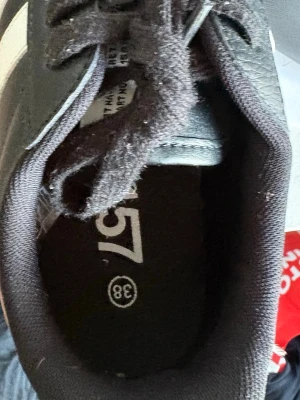 Svarta sneakers från 157, strl 38 - Svarta sneakers från 157 med vit sidodetalj och bruna gummisulor. Skorna har klassisk låg profil, snörning och ovandel i syntetmaterial. Perfekta för en avslappnad streetstil.