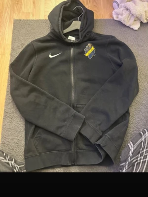 Svart AIK hoodie  - Svart hoodie med dragkedja från Nike, prydd med AIK:s klubbmärke på bröstet. Klassisk huva och två fickor framtill. Perfekt för dig som vill representera AIK på ett stilrent sätt. Tillverkad i mjukt bomullsmaterial för skön känsla.