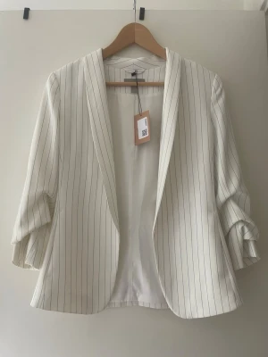 Vit randig kavaj från H&M - Snygg vit kavaj från H&M med tunna svarta ränder. Oversized passform med uppvikta ärmar och öppen front utan knappar. Tillverkad i ett lätt tyg som ger en stilren och modern look. Perfekt för att lyfta vilken outfit som helst.