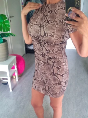 Kort klänning med ormmönster - Säljer en snygg kort klänning med trendigt ormmönster i beige och bruna toner. Klänningen har hög hals, korta ärmar och en tight passform som framhäver figuren. Perfekt för dig som vill sticka ut med en cool look.