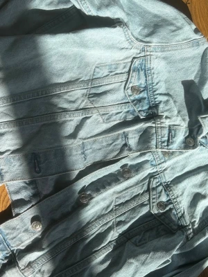 Ljusblå klassisk jeansjacka - Snygg ljusblå jeansjacka med silverfärgade knappar och två bröstfickor med lock. Jackan har en tidlös design och raka sömmar som ger en clean look. Perfekt att slänga över en hoodie eller t-shirt för en avslappnad stil.