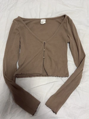 Beige ribbad croppad topp - Trendig beige croppad topp med lång ärm och v-ringning. Toppen har ribbad struktur, knappar framtill och lätt vågig kant nedtill. Perfekt för att styla med höga jeans eller kjol.