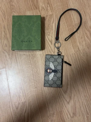 GG plånbok med bi och handledsrem - Snygg plånbok från GG med klassiskt GG-mönster och en stor bi-detalj på framsidan. Plånboken har en avtagbar handledsrem i svart med gröna och röda ränder samt dragkedja. Perfekt för dig som vill ha något unikt och trendigt.