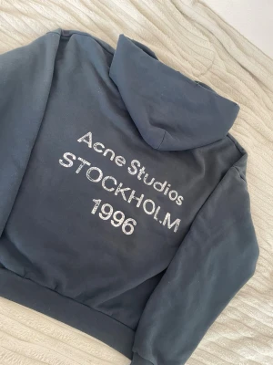 Svart hoodie Acne Studios Stockholm - Snygg svart hoodie från Acne Studios med stor huva och tryck på ryggen: 'Acne Studios STOCKHOLM 1996' i vitt. Klassisk passform med ribbade muddar och mjukt material, perfekt för en chill och trendig look.